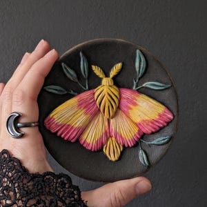 Puede incluir: Una placa decorativa circular de color negro con una polilla en relieve de textura marcada y alas en tonos amarillo y rosa vibrantes. La polilla está rodeada por pequeñas hojas esculpidas en color verde azulado. Acabado mate.