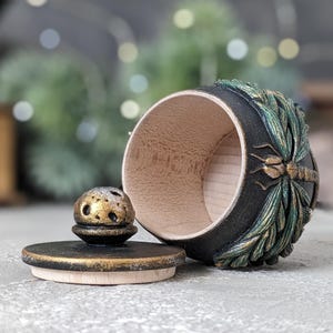 May include: A small, round wooden container with a black exterior and a natural light wood interior. The side features a relief carving of a dragonfly with green and gold accents. A matching black lid with a gold-dusted knob sits beside it.