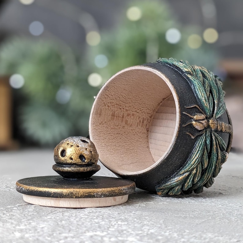 May include: A small, round wooden container with a black exterior and a natural light wood interior. The side features a relief carving of a dragonfly with green and gold accents. A matching black lid with a gold-dusted knob sits beside it.