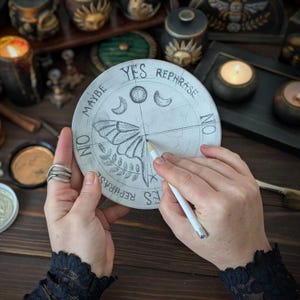 May include: A circular divination board featuring a hand-drawn moth, moon phases, and the words YES, NO, MAYBE, and REPHRASE. The board is light-colored with dark ink illustrations and is being marked with a white pencil at the center point.