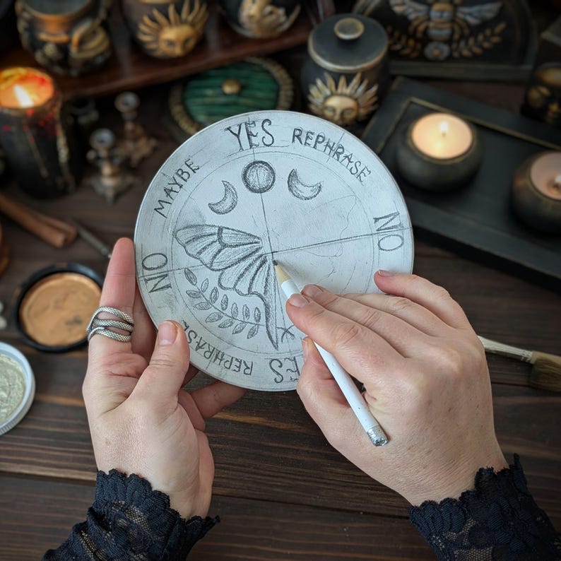 May include: A circular divination board featuring a hand-drawn moth, moon phases, and the words YES, NO, MAYBE, and REPHRASE. The board is light-colored with dark ink illustrations and is being marked with a white pencil at the center point.
