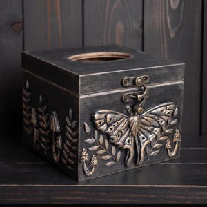Puede incluir: Una caja de pa&ntilde;uelos cuadrada de madera con un acabado oscuro y desgastado. El frente presenta un emblema met&aacute;lico de polilla en relieve rodeado de motivos de helechos y setas. Un cierre de metal color lat&oacute;n asegura la tapa abatible.