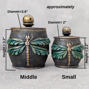 May include: Two dark gray lidded jars with gold and green dragonfly motifs. The larger jar is 3.2 inches high with a 2.6-inch diameter. The smaller jar is 2.2 inches high with a 2-inch diameter. Both feature round lids with small knobs.