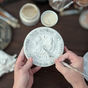 May include: A circular off-white clay divination board featuring a central embossed moth, moon phases, and leaf sprigs. The text YES, NO, MAYBE, and REPHRASE is inscribed around the perimeter. A thin brush is applying paint to the moth detail.