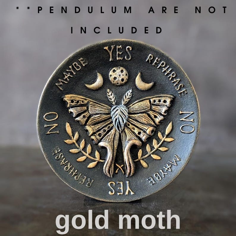 May include: A circular dark metal divination board featuring a central gold-toned moth, a moon, and a sun. The text reads YES, NO, MAYBE, and REPHRASE. The top text states PENDULUM ARE NOT INCLUDED and the bottom text reads gold moth.