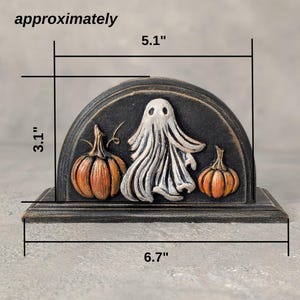 May include: A semi-circular black decorative stand featuring a raised white ghost figure flanked by two orange pumpkins. The piece measures approximately 13 cm in height, 7.9 cm in depth, and 17 cm in width as indicated by the text.