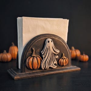 May include: A dark-finished wooden napkin holder featuring a semi-circular front panel decorated with a carved, raised white ghost figure flanked by two orange pumpkins. The holder contains a stack of plain, light beige paper napkins.