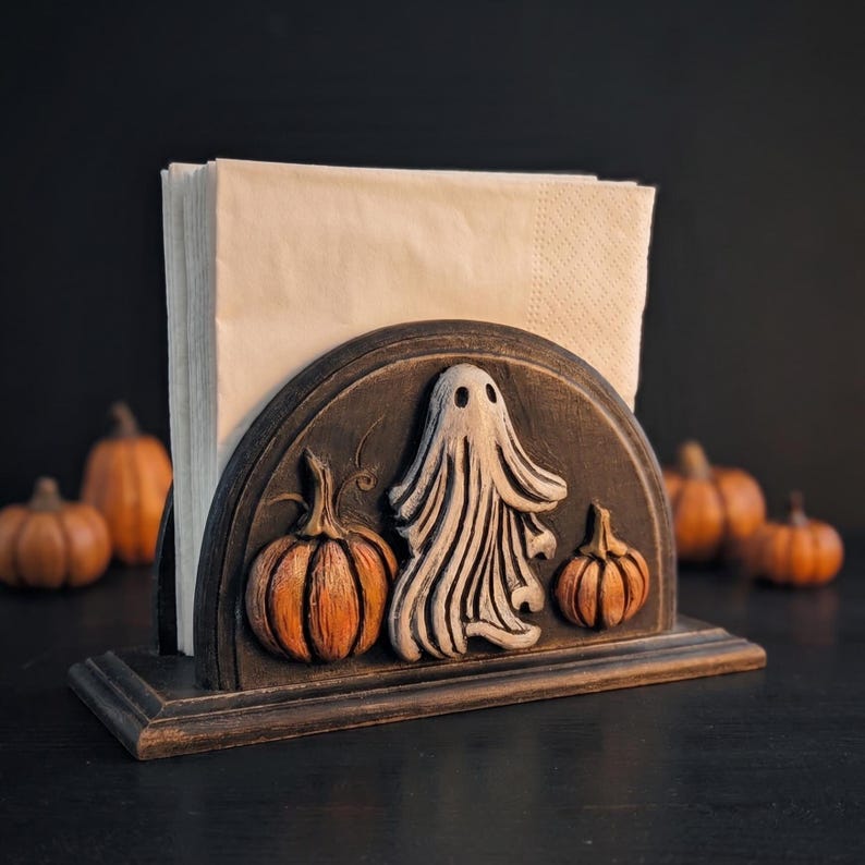 May include: A dark-finished wooden napkin holder featuring a semi-circular front panel decorated with a carved, raised white ghost figure flanked by two orange pumpkins. The holder contains a stack of plain, light beige paper napkins.