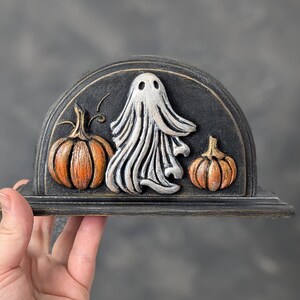May include: A semi-circular black napkin holder featuring a raised relief design of a central white ghost flanked by two orange pumpkins. The piece has a distressed finish with visible brush strokes and a flat base for stability on a surface.