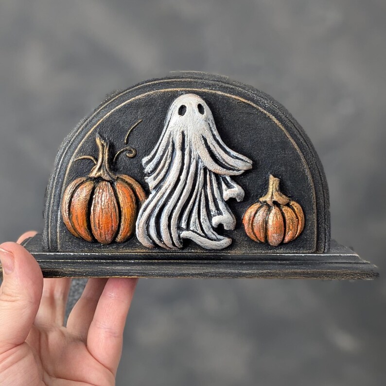 May include: A semi-circular black napkin holder featuring a raised relief design of a central white ghost flanked by two orange pumpkins. The piece has a distressed finish with visible brush strokes and a flat base for stability on a surface.