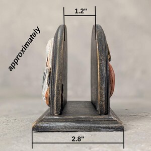 May include: A dark, distressed-finish wooden napkin holder with two upright panels and a flat base. The interior width measures 1.2 inches, and the total base width measures 2.8 inches, as indicated by the text and lines on the image.