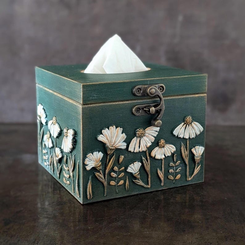 May include: A teal wooden tissue box with a hinged lid and metal clasp. The box is decorated with white and gold floral relief carvings. A stack of white tissues is visible at the top.