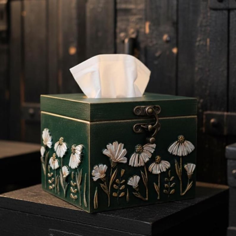May include: A dark green wooden tissue box with a hinged lid and a decorative floral design. White tissues are visible at the top. The box is square-shaped and sits on a dark surface.