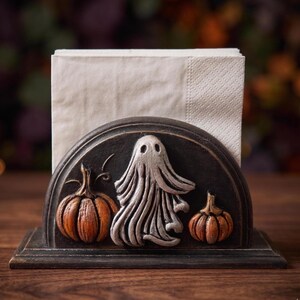 May include: A black semi-circular napkin holder featuring a raised relief design of a central silver ghost flanked by two orange pumpkins. The holder is constructed with a flat base and holds a stack of plain white paper napkins upright.