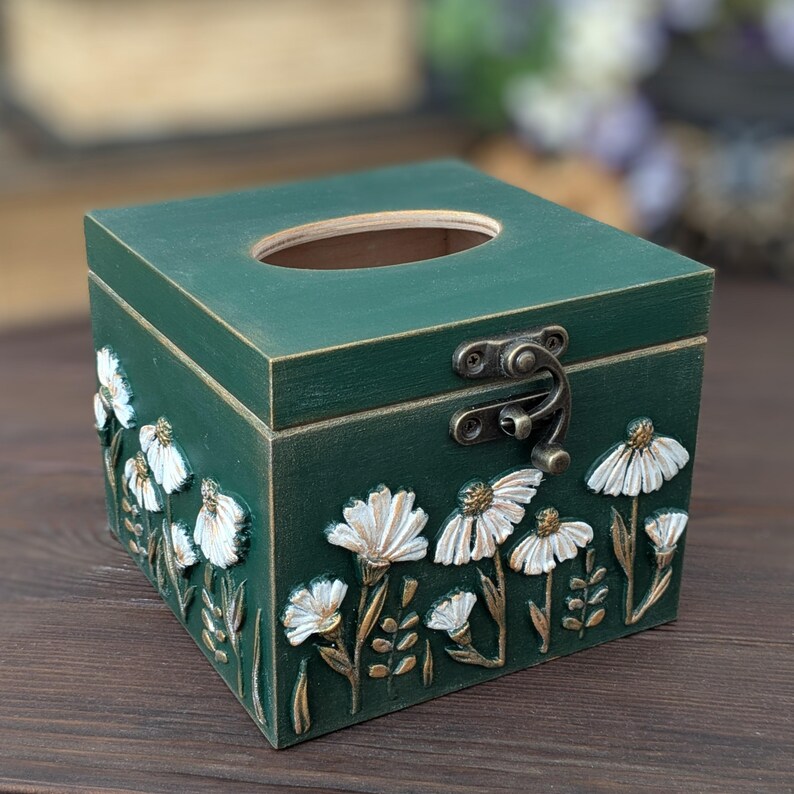 May include: A square, dark green wooden tissue box with a hinged lid and a decorative floral design. The box features white and gold embossed flowers and a metal clasp. The top has an oval opening for tissue dispensing.