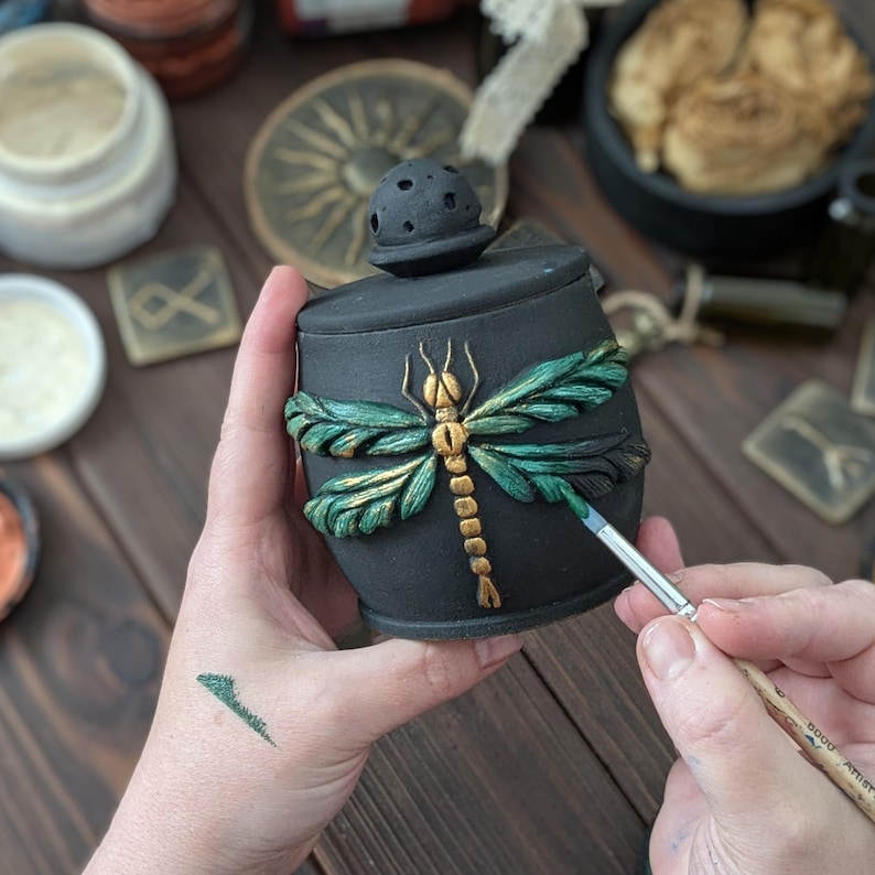 May include: A matte black ceramic jar with a lid featuring a sculpted dragonfly with metallic green and gold wings and body. The jar is being painted with a fine-tipped brush, highlighting the textured details of the insect relief design.