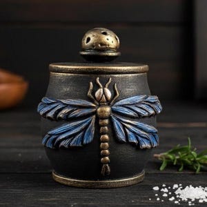 May include: Black and bronze salt shaker with a dragonfly design. The salt shaker has a cylindrical shape with a lid featuring small holes. The dragonfly is detailed with blue wings and is centered on the body of the shaker.