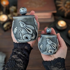 Puede incluir: Dos tarros decorativos de color gris oscuro con tapas, con un relieve en relieve de un conejo y espigas de trigo. Los tarros se sostienen en las manos, mostrando la artesan&iacute;a y el dise&ntilde;o detallados. Las tapas tienen un peque&ntilde;o pomo redondo.