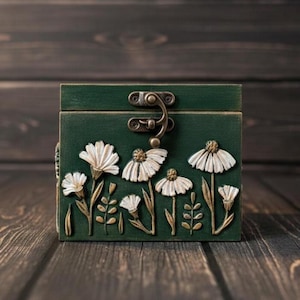 May include: A green wooden box with a metal clasp, adorned with white daisy and leaf relief carvings. The box is set against a dark wood background, showcasing the floral design and vintage aesthetic.