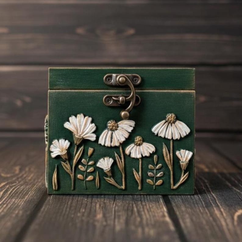 May include: A green wooden box with a metal clasp, adorned with white daisy and leaf relief carvings. The box is set against a dark wood background, showcasing the floral design and vintage aesthetic.