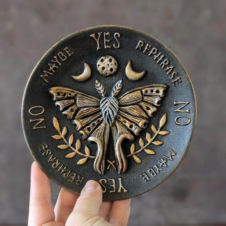 May include: A circular black and gold plaque featuring a central moth illustration surrounded by celestial symbols including a moon and stars. The text YES, NO, MAYBE, and REPHRASE is embossed around the perimeter in a circular arrangement.