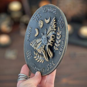 May include: An oval-shaped plaque with a dark, weathered finish featuring a central moth relief. The surface is embossed with celestial symbols including a moon and stars, along with the words YES, NO, MAYBE, and REPHRASE arranged around the moth.
