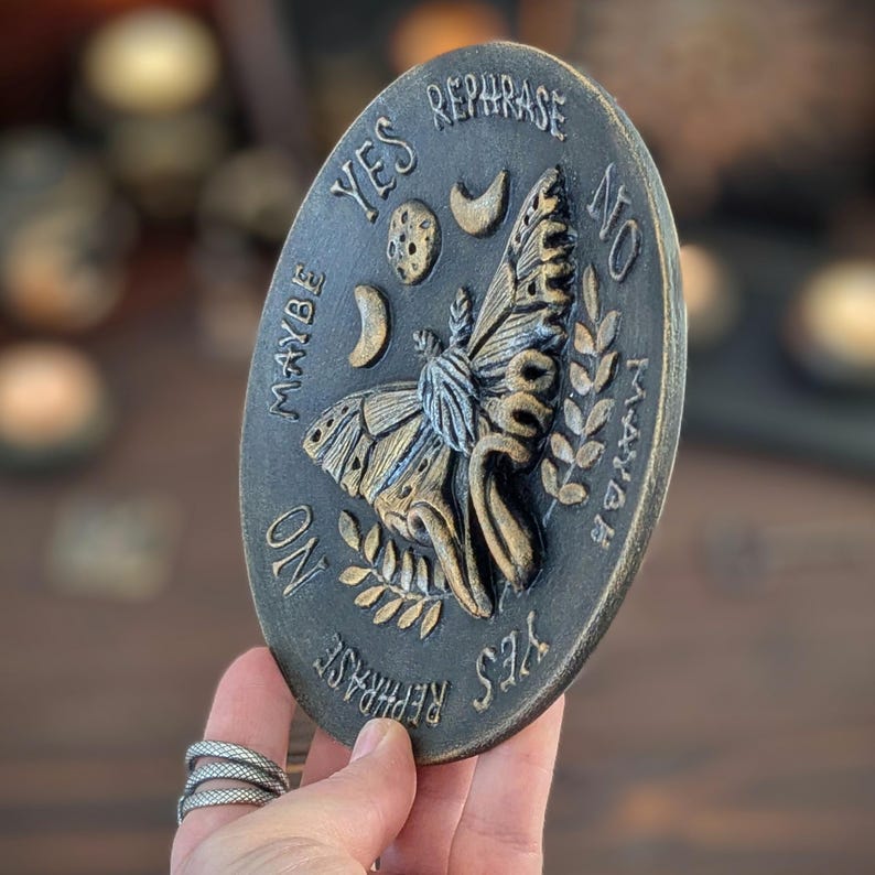 May include: An oval-shaped plaque with a dark, weathered finish featuring a central moth relief. The surface is embossed with celestial symbols including a moon and stars, along with the words YES, NO, MAYBE, and REPHRASE arranged around the moth.