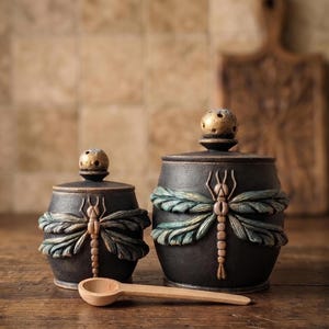 May include: Two dark charcoal ceramic canisters with lids, each featuring a raised, sculpted dragonfly with teal and bronze wings. The lids are topped with small, textured gold spheres. A light-colored wooden spoon rests horizontally in front.