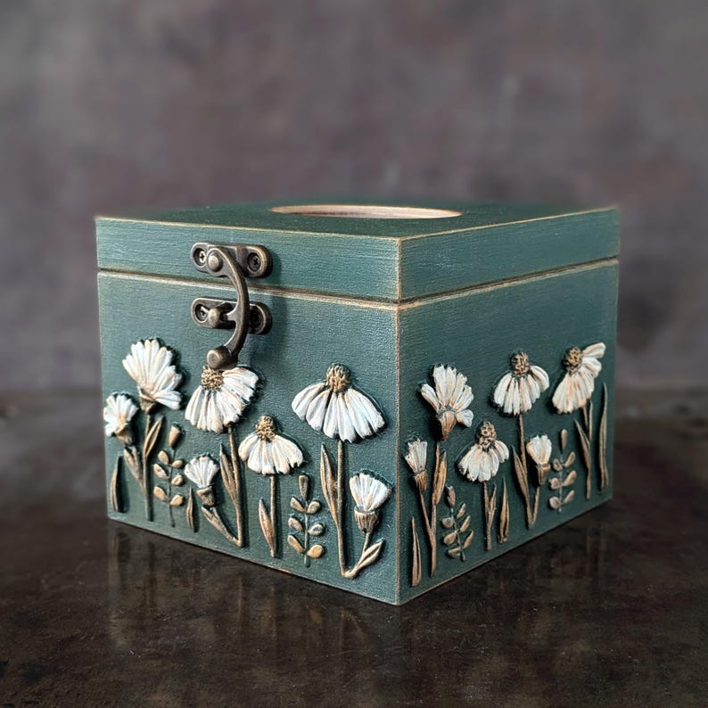 May include: A teal wooden box with a hinged lid and metal clasp. The box is decorated with white and gold floral relief carvings. The box has a square shape and a hole in the top.