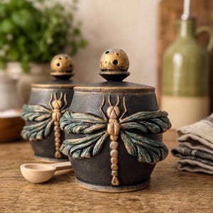 May include: Two round black ceramic canisters with lids, each featuring a raised gold and teal dragonfly relief design. The lids have a small spherical knob with perforated holes. A small wooden spoon rests in front of the canisters.