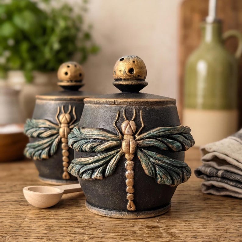 May include: Two round black ceramic canisters with lids, each featuring a raised gold and teal dragonfly relief design. The lids have a small spherical knob with perforated holes. A small wooden spoon rests in front of the canisters.