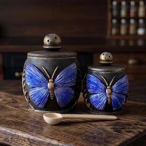 May include: Two black ceramic jars with lids, each featuring a raised blue butterfly design with gold accents. The jars have rounded bodies and small knob handles on the lids. A small, light-coloured wooden spoon is placed horizontally in front of them.