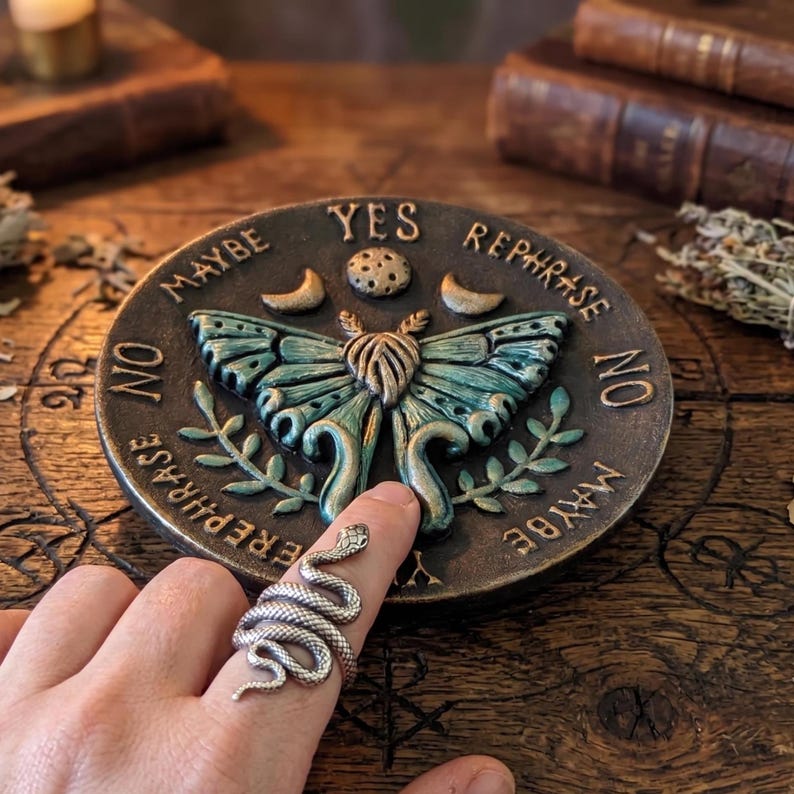May include: A round dark brown plaque featuring a central teal and bronze moth, a moon, and a sun. The text YES, NO, MAYBE, and REPHRASE is embossed around the edge. A silver-toned snake ring is worn on a finger pointing toward the moth design.