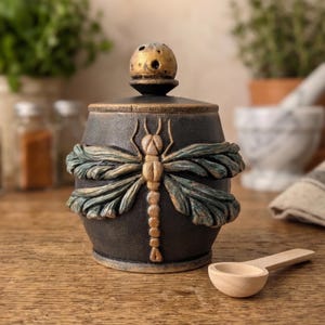 May include: A dark charcoal-colored ceramic jar with a matching lid featuring a textured, raised dragonfly relief in muted gold and teal tones. The lid has a spherical knob, and a small, light-colored wooden spoon rests on the surface nearby.