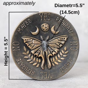May include: A circular bronze-toned plaque featuring a central moth, moon, and sun design. The text reads YES, NO, MAYBE, and REPHRASE. It measures 5.5 inches in height and 5.5 inches in diameter. The surface has a weathered, textured finish.