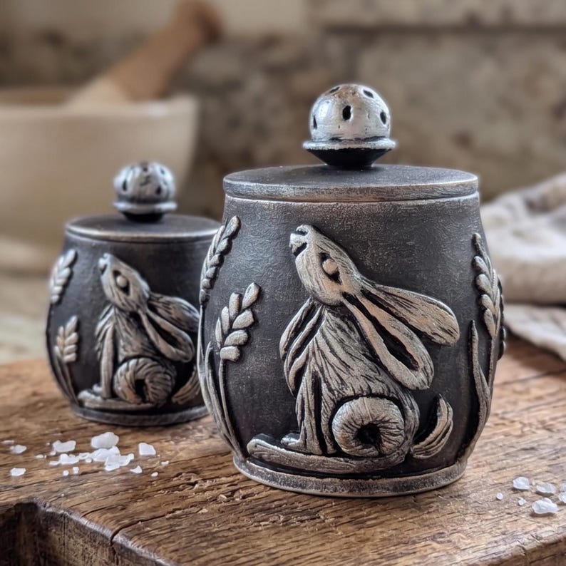 Puede incluir: Dos saleros y pimenteros de color gris oscuro con un dise&ntilde;o de conejo y trigo. Los saleros tienen un relieve de conejo plateado y espigas de trigo. Las tapas tienen un peque&ntilde;o pomo redondo plateado con agujeros. Los saleros est&aacute;n sobre una superficie de madera.
