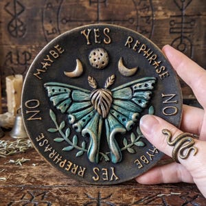 May include: A round, dark bronze-toned divination board featuring a central teal and gold moth, a moon, and stars. The text reads YES, NO, MAYBE, and REPHRASE. The board is held by a hand wearing a gold-toned snake ring.