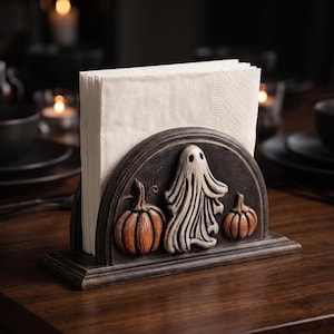 May include: A dark-toned, semi-circular napkin holder featuring a raised, sculpted design of a central white ghost flanked by two orange pumpkins. The holder is constructed with a flat base and a vertical slot currently filled with white paper napkins.