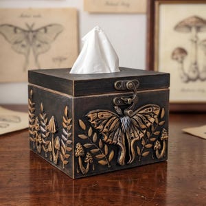 Puede incluir: Una caja de pa&ntilde;uelos cuadrada de color marr&oacute;n oscuro con tapa abatible y cierre met&aacute;lico. El exterior presenta detalles dorados en relieve de una gran polilla, varios hongos y ramas con hojas. Un pa&ntilde;uelo blanco asoma por arriba.