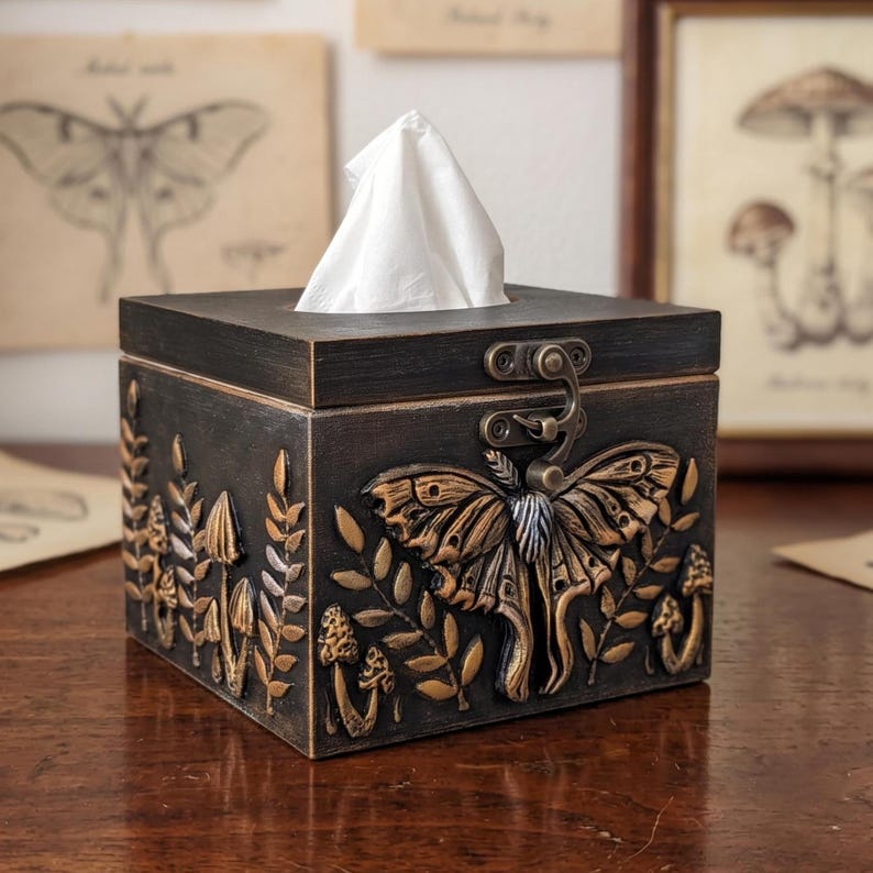 Puede incluir: Una caja de pa&ntilde;uelos cuadrada de color marr&oacute;n oscuro con tapa abatible y cierre met&aacute;lico. El exterior presenta detalles dorados en relieve de una gran polilla, varios hongos y ramas con hojas. Un pa&ntilde;uelo blanco asoma por arriba.