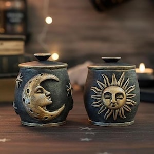 May include: Two small, dark gray cylindrical jars with matching lids. One jar features a raised, gold-toned crescent moon with a face and small stars. The other jar displays a raised, gold-toned sun with a face and radiating rays.