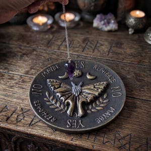 May include: A circular dark metal divination board featuring a central moth relief, crescent moons, and the words YES, NO, MAYBE, and REPHRASE. A purple amethyst crystal pendulum hangs from a silver chain above the board.