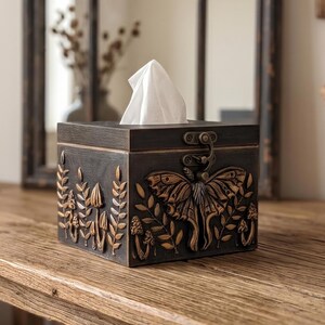 Puede incluir: Una caja cuadrada de color marr&oacute;n oscuro para pa&ntilde;uelos con tapa abatible y cierre met&aacute;lico. El exterior presenta detalles dorados en relieve que incluyen una gran polilla central, varios hongos y ramas de helecho en los paneles.