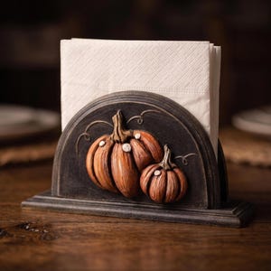 Puede incluir: Un servilletero negro semicircular con un diseño en relieve de dos calabazas naranjas con tallos marrones y pequeños detalles blancos. El soporte tiene una base plana y contiene una pila de servilletas de papel blancas plegadas.
