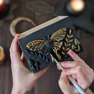 Puede incluir: Una caja rectangular negra con una escultura en relieve tridimensional de una polilla dorada y negra en la parte frontal. El dise&ntilde;o incluye ramas doradas en relieve y peque&ntilde;as formas de setas. Un pincel aplica pintura dorada sobre la polilla.