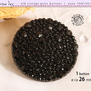 May include: A black, vintage glass button, approximately 26 mm in diameter, covered in small beads and larger, faceted accents. The button is round and features text that reads "rosa day... old vintage glass buttons | anno 1940/50/60".