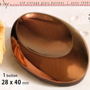 Könnte beinhalten: Ein ovaler Vintage-Glasknopf mit einer reflektierenden, metallisch kupferfarbenen Oberfläche. Der Knopf hat eine glatte, gewölbte Form und ist mit dem Text 1 button je ca 28 x 40 mm beschriftet, der die Maße in Millimetern angibt.