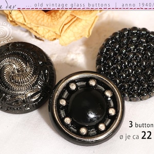Könnte beinhalten: Drei antike schwarze Glas Knöpfe mit einzigartigen Designs. Ein Knopf hat ein Spiralmuster, ein anderer eine strukturierte Oberfläche, und der dritte einen zentralen schwarzen Kreis mit silbernen Akzenten. Jeder Knopf hat einen Durchmesser von ca. 22 mm.