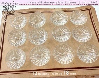 May include: Twelve clear glass buttons with a swirl design, arranged in a grid on a brown paper card. The card has the text "12 buttons øje ca 18 mm" printed on it.