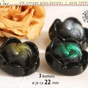 Könnte beinhalten: Drei schwarze Glasknöpfe in Blumenform mit irisierenden Zentren in Grün- und Goldtönen. Die Knöpfe haben eine strukturierte Oberfläche und einen Durchmesser von ca. 22 mm. Der Text auf dem Bild lautet: rosa day, old vintage glass buttons, anno 1940/50/60, design by karin, 3 buttons, ø je ca 22 mm.